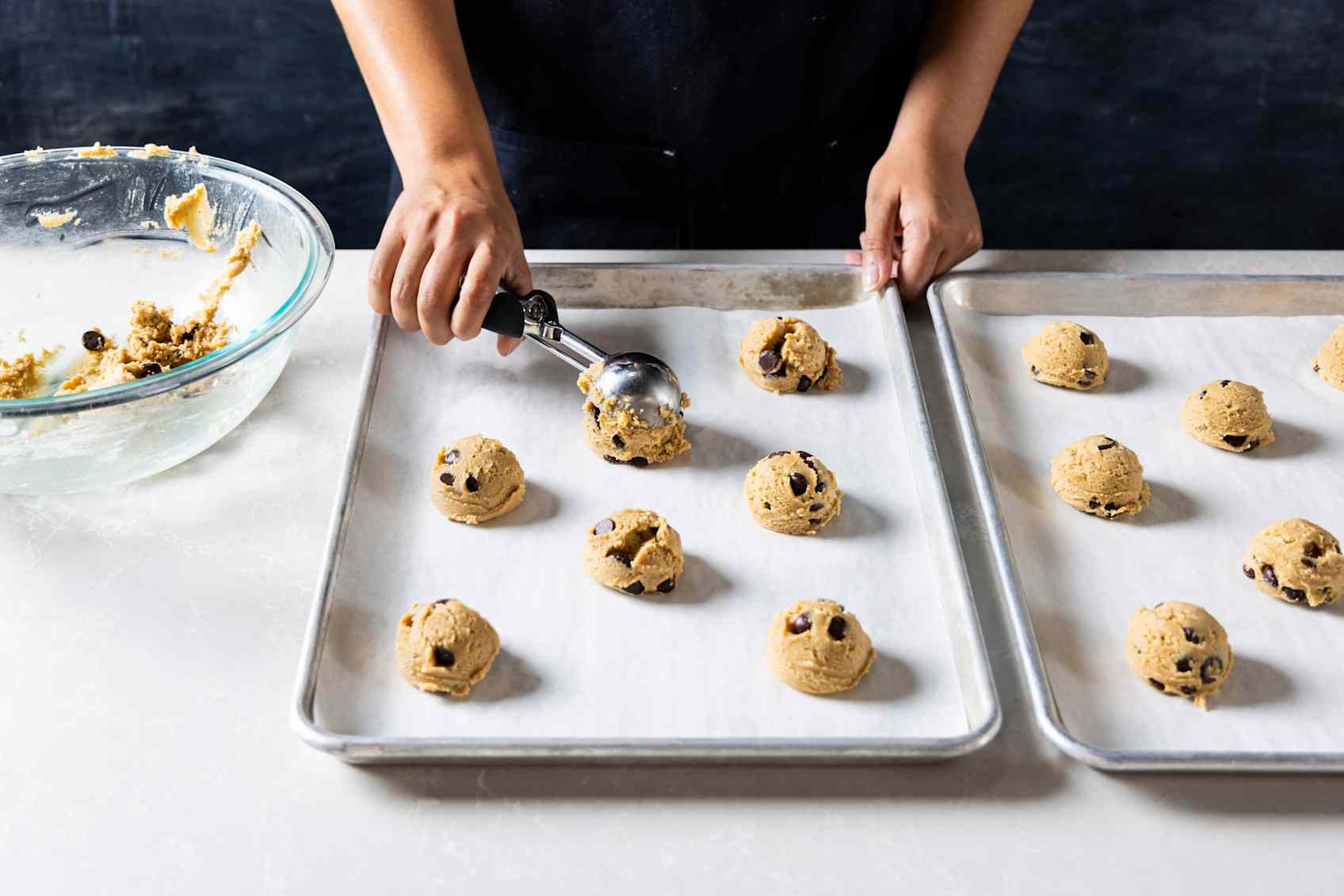 Hands dispensing a ball of cookie dough onto a baking sheet lined with parchment paper. 